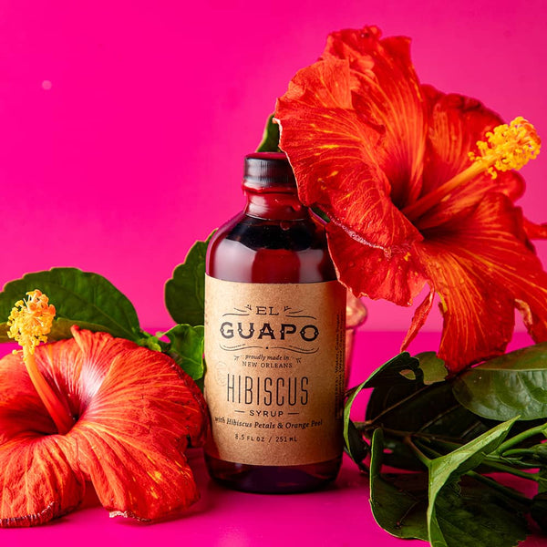 Bottle of El Guapo Hibiscus Syrup with red hibiscus flowers on a pink background